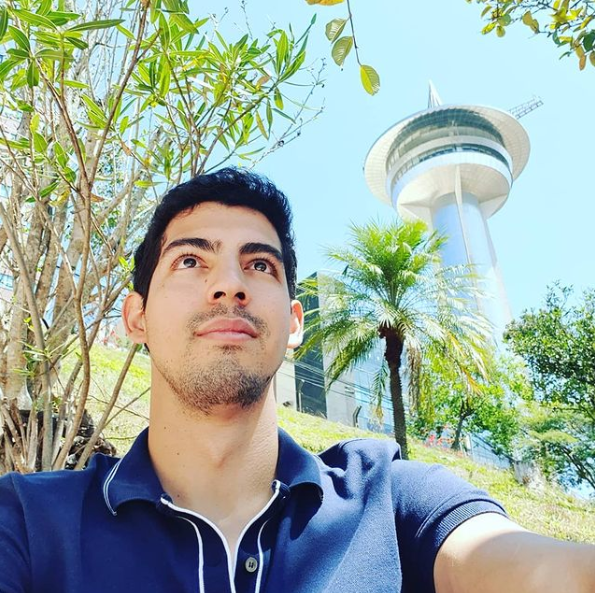 Antonio Fernandes ao ar livre, com árvores e uma torre ao fundo